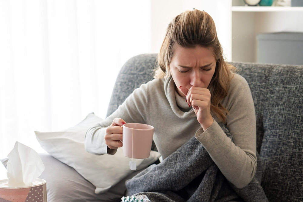 A women caughing while holding a cup of tea in other hand