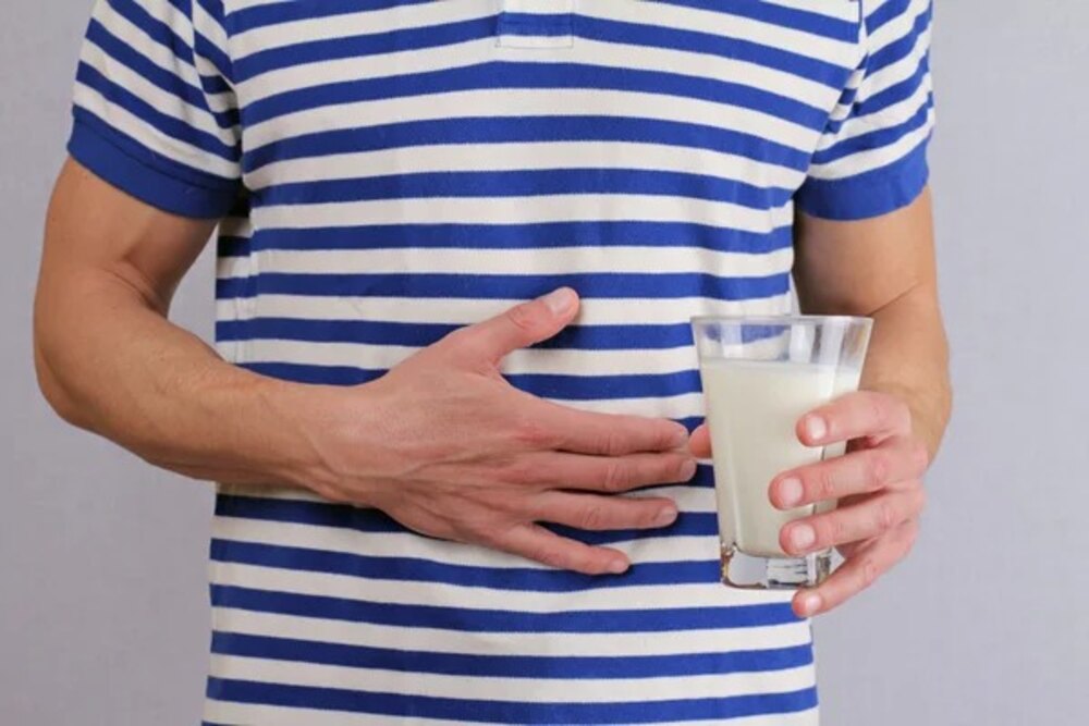 A person holding a glass of milk in one hand and placing the other on their stomach, depicting lactose intolerance and digestive discomfort