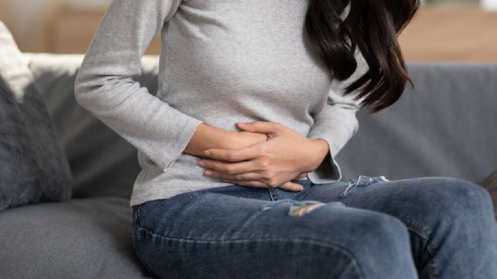 A woman sitting indoors holds her lower abdomen with both hands, showing sign of UTI