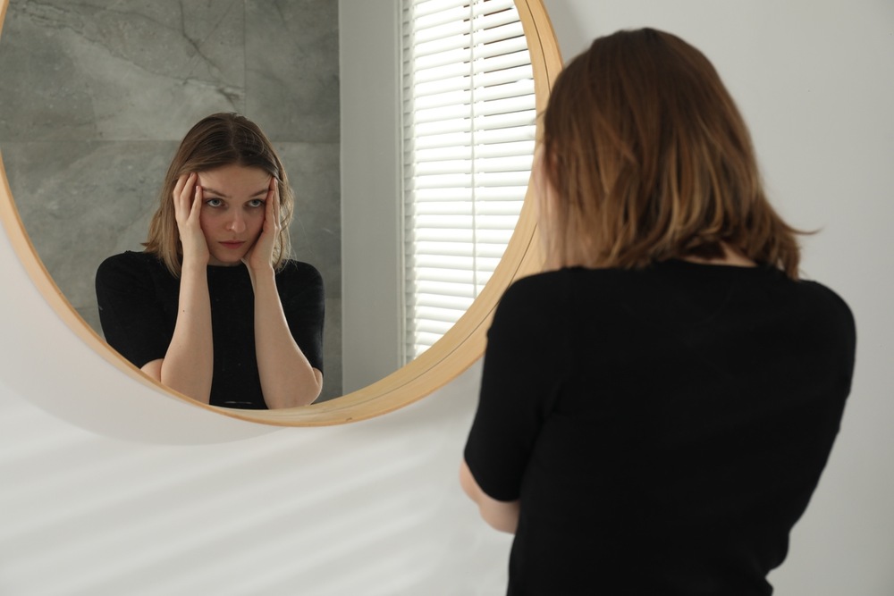 Woman looking anxiously into a mirror while examining her body, illustrating concerns about perceived physical flaws associated with body dysmorphic disorder