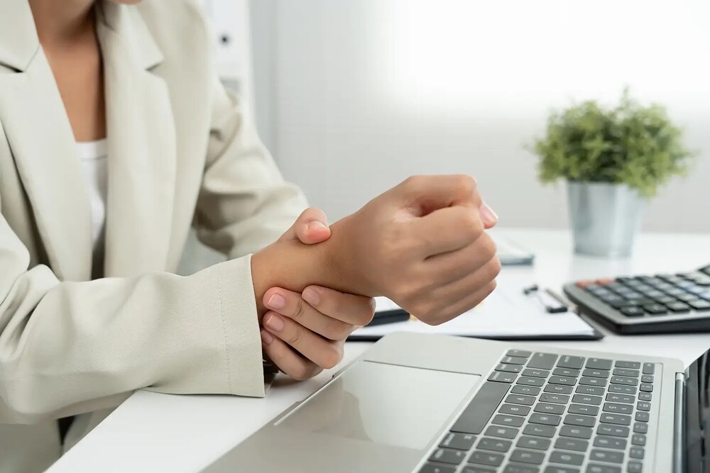 A woman sitting in front of a laptop gently holds her wrist with a painful expression, illustrating carpal tunnel syndrome