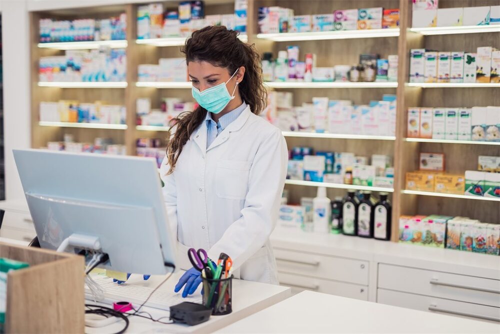 Pharmacist working at a computer beside medicine shelves, illustrating a report on insulin supply disruption in Pakistan.