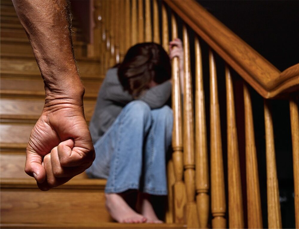 Child sitting on wooden stairs with their face hidden and an adult’s clenched fist in the foreground, illustrating physical and psychological violence against children.