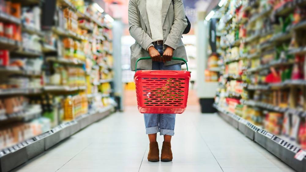 A woman standing in a grocery store holding a shopping basket, comparing packaged food products and checking their shelf-life dates.
